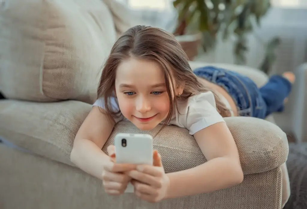 A young girl with long hair, wearing a white t-shirt and jeans, is lying on a beige couch. She smiles while exploring KidSmartⓇ Apps on her smartphone, the best kid-safe messaging choice. A blurred plant subtly decorates the background.