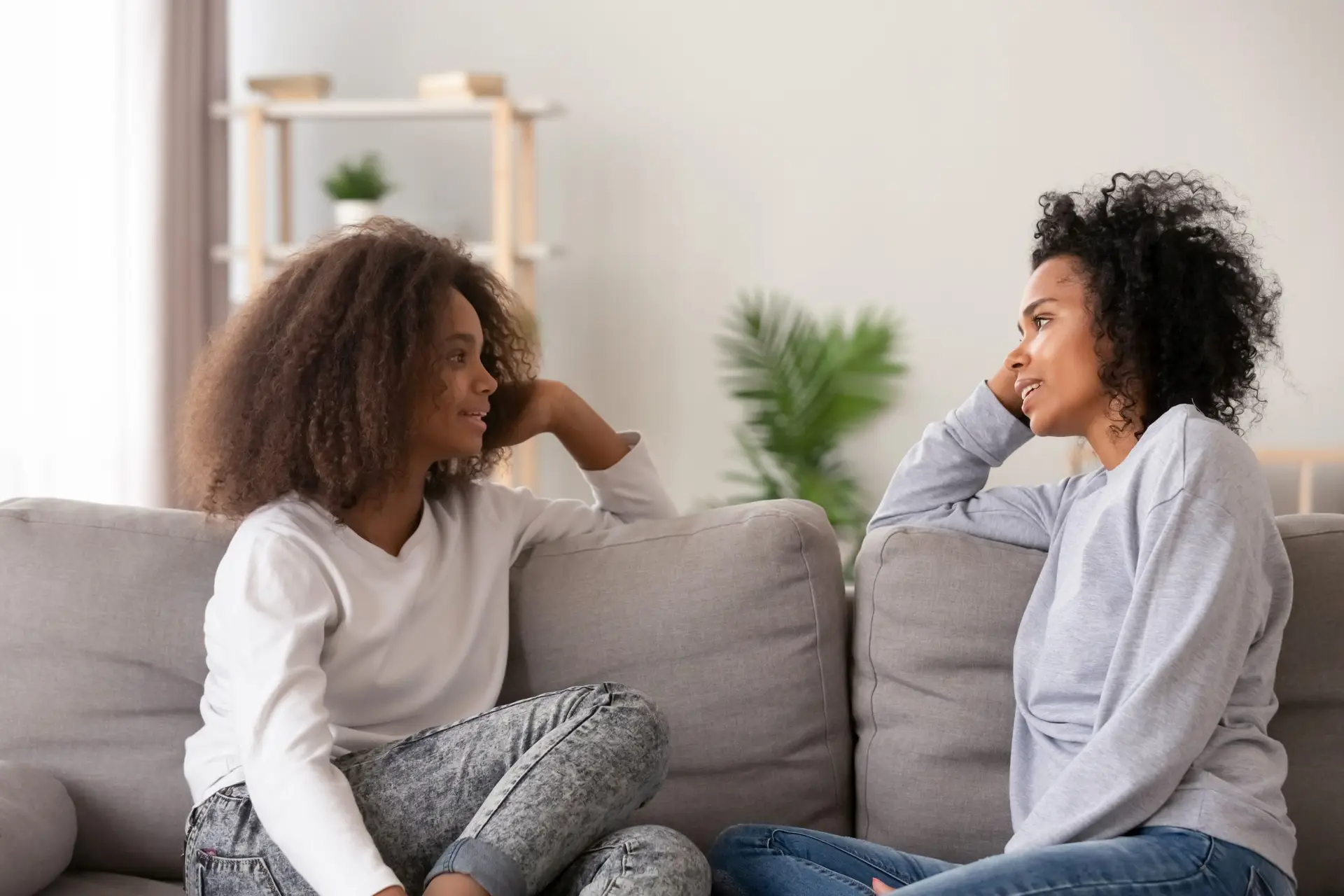 Two women with curly hair sit on a sofa facing each other, casually discussing parenting topics like "Is It Okay to Take Your Child’s Phone Away?" amid the cozy setting of a neutral-toned room with a potted plant adding to the tranquil atmosphere.