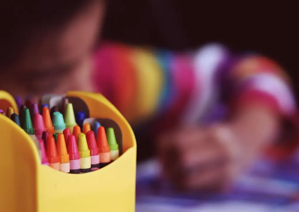 A box of colorful crayons is in focus in the foreground. In the blurred background, a child in a rainbow-striped shirt joyfully engages in summer art for kids, drawing on paper.