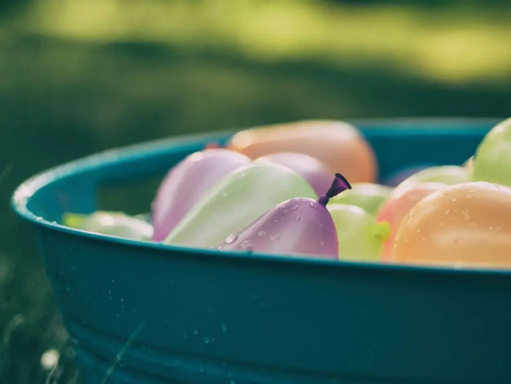 A blue tub filled with colorful water balloons, glistening with droplets of water, sits on the grass—perfect for water games for kids. The blurred green background suggests an outdoor setting, likely a sunny day in a park or backyard.