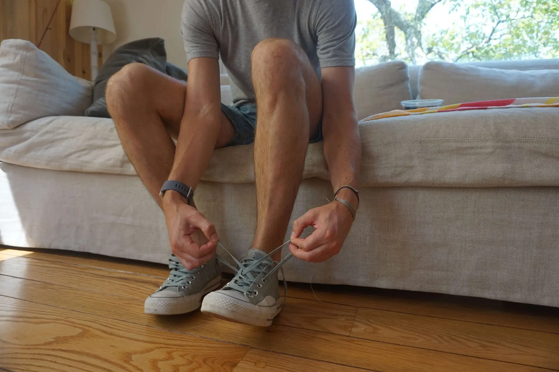 A person wearing a gray t-shirt and shorts sits on a beige couch, demonstrating how to teach kids to tie shoes by carefully tying one of their gray sneakers. The room, with its wooden flooring and lamp in the background, is softly illuminated as sunlight filters through a window.