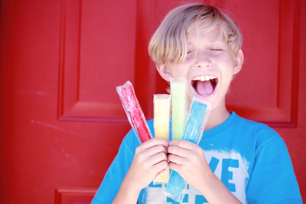 A child with light hair is smiling widely, showcasing their summer snacks for kids: four colorful popsicles—red, orange, yellow, and green—against a bright red door. The child is wearing a blue T-shirt with white text.