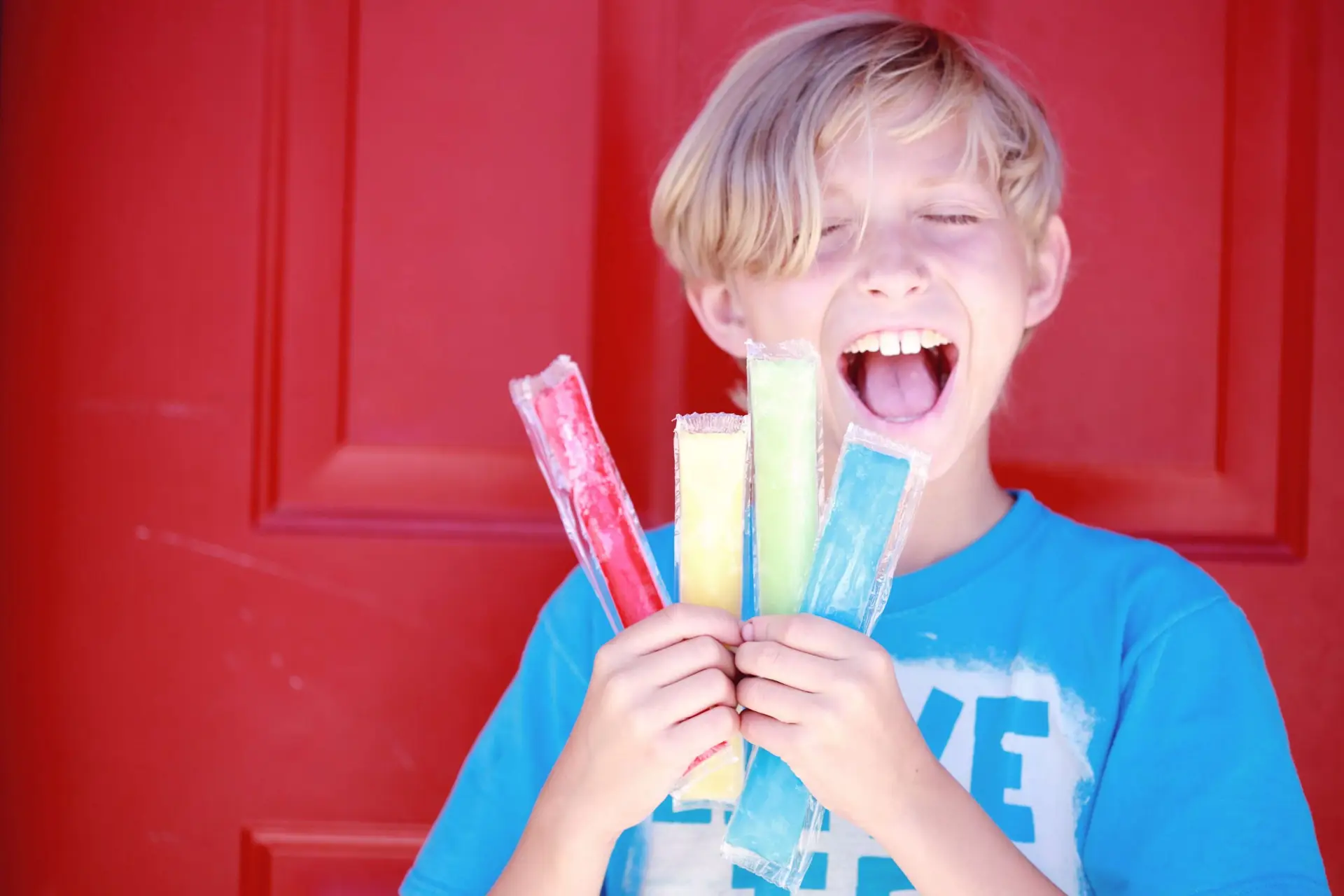 A child with light hair is smiling widely, showcasing their summer snacks for kids: four colorful popsicles—red, orange, yellow, and green—against a bright red door. The child is wearing a blue T-shirt with white text.