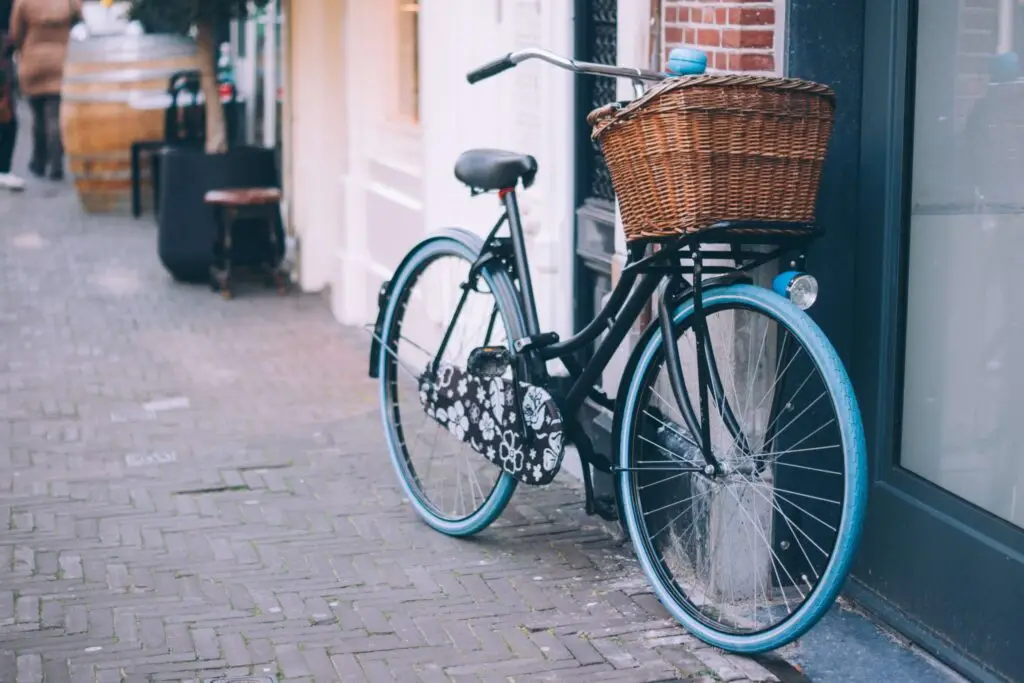 A black bicycle, equipped with a charming kids bike basket and blue tires, rests on a brick-paved street in front of a building with a glass door. Nearby, a wooden barrel and parts of another storefront enhance the quaint scene.