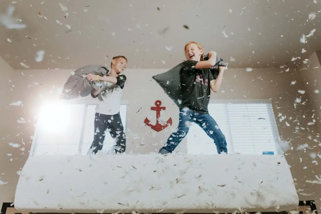 Two children joyfully have a pillow fight on a bed, feathers flying around as they embrace growth mindset activities for kids. They're smiling in a bright room, with a red anchor decoration on the wall behind them.