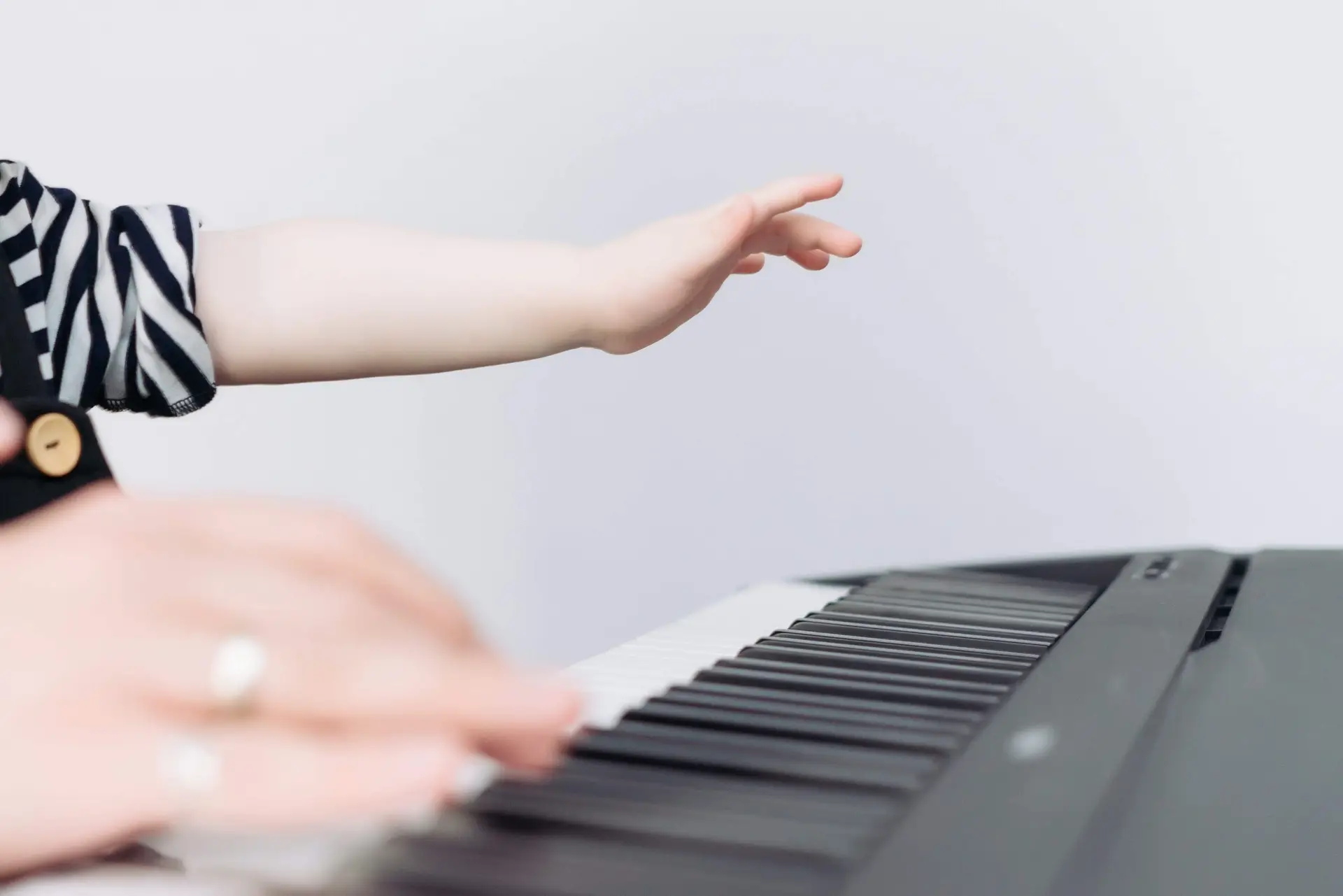 A child's hand reaches over the keys of a black electronic keyboard, striped shirt partially visible. Beside it, another hand plays on. Set against a plain light background, it's as if notes from piano books for kids are coming to life in a vivid duet.