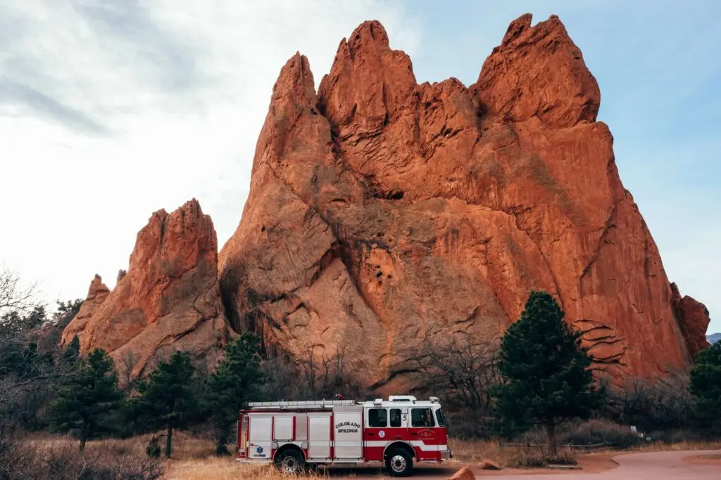 A fire truck is parked on a road in front of towering red rock formations with rugged peaks. Sparse trees surround the area, and the sky is cloudy, creating a dramatic backdrop—an ideal setting for teaching fire safety tips for kids amidst nature's grandeur.