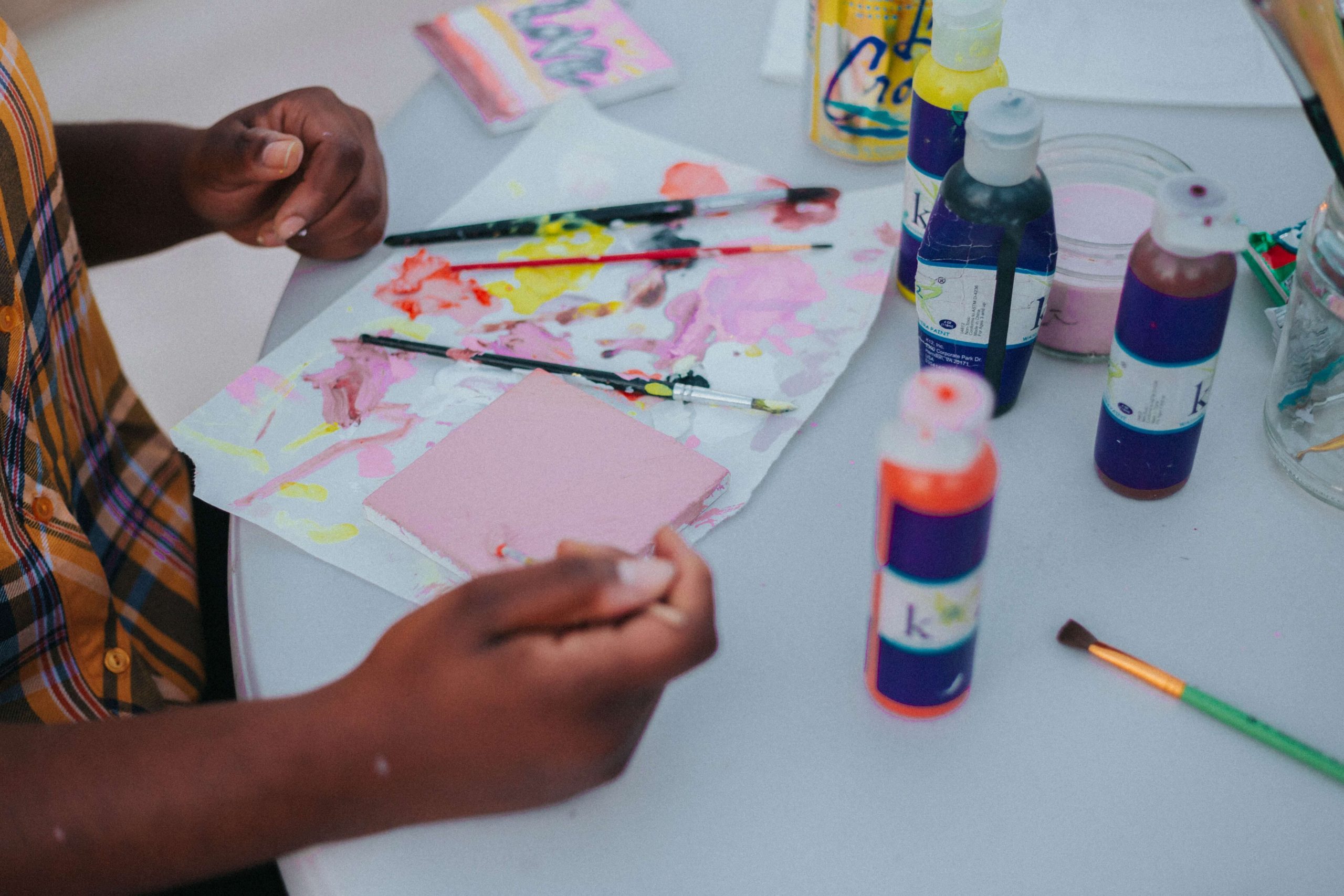 A person is painting at a table, surrounded by several brushes, paint bottles, and paper, evoking the lively spirit of a paint party for kids. Their hands are busy on a small pink canvas. A can of La Croix and colorful paints nearby enhance the vibrant workspace.