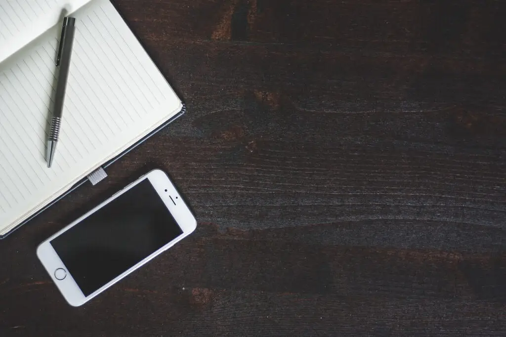 An open notebook with a pen rests on a dark wooden surface, inviting thoughts to flow, while a smartphone nearby serves as a portal for staying connected and discovering how Life360 works in harmonizing family life. The scene conveys a simple, organized workspace setup.