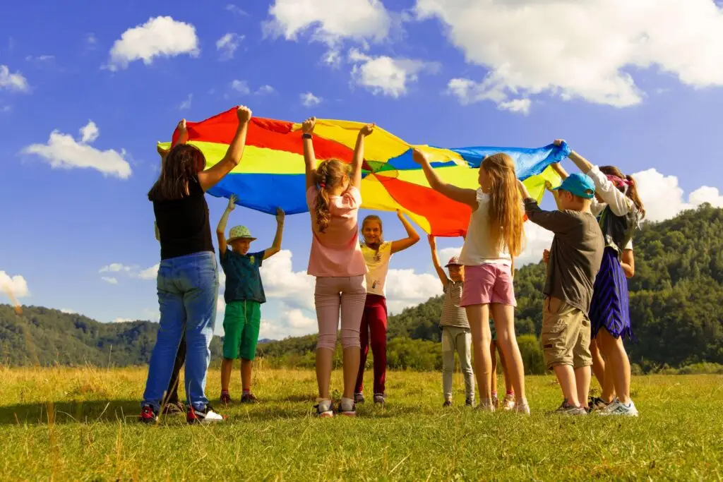 A group of children stands in a grassy field, enjoying summer camp activities for kids as they hold up a colorful parachute under a blue sky with clouds. Forested hills are visible in the background, adding to the joyful outdoor setting.