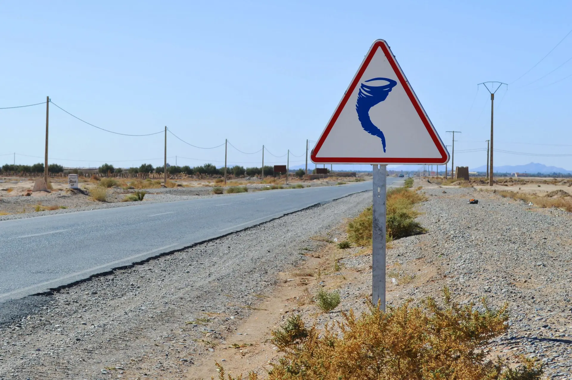 A triangular warning sign with a tornado symbol stands beside an empty desert road under a clear blue sky, reminding us of the importance of tornado safety for kids. Sparse vegetation and utility poles line the road, creating an eerie yet serene landscape.