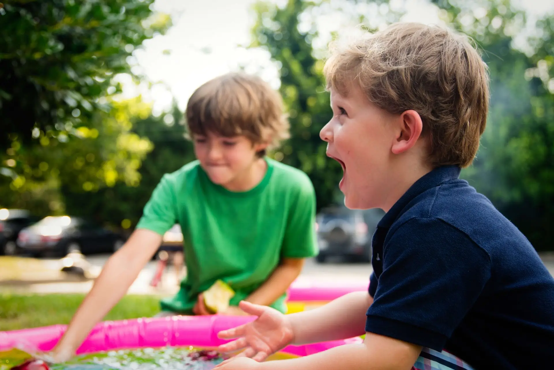 Two young boys play joyfully outdoors near a pink inflatable pool, amidst the lively buzz of outdoor party games for kids. One boy with messy brown hair reaches into the water, while the other, wearing a navy shirt, looks excitedly to the side. Green trees and parked cars are visible in the background.