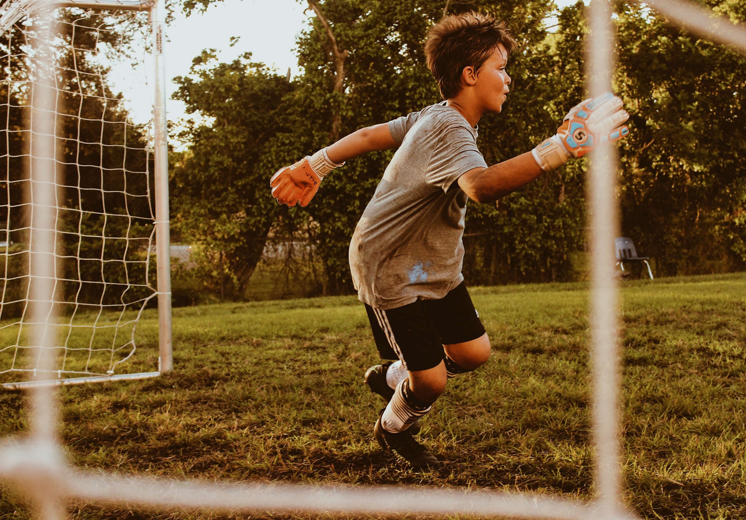 A young soccer goalie in gloves and a gray shirt defends the goal during a fun soccer game for kids. The sunlit field, with green grass and trees in the background, adds to the lively atmosphere, while the goal net stands partially visible in the foreground.