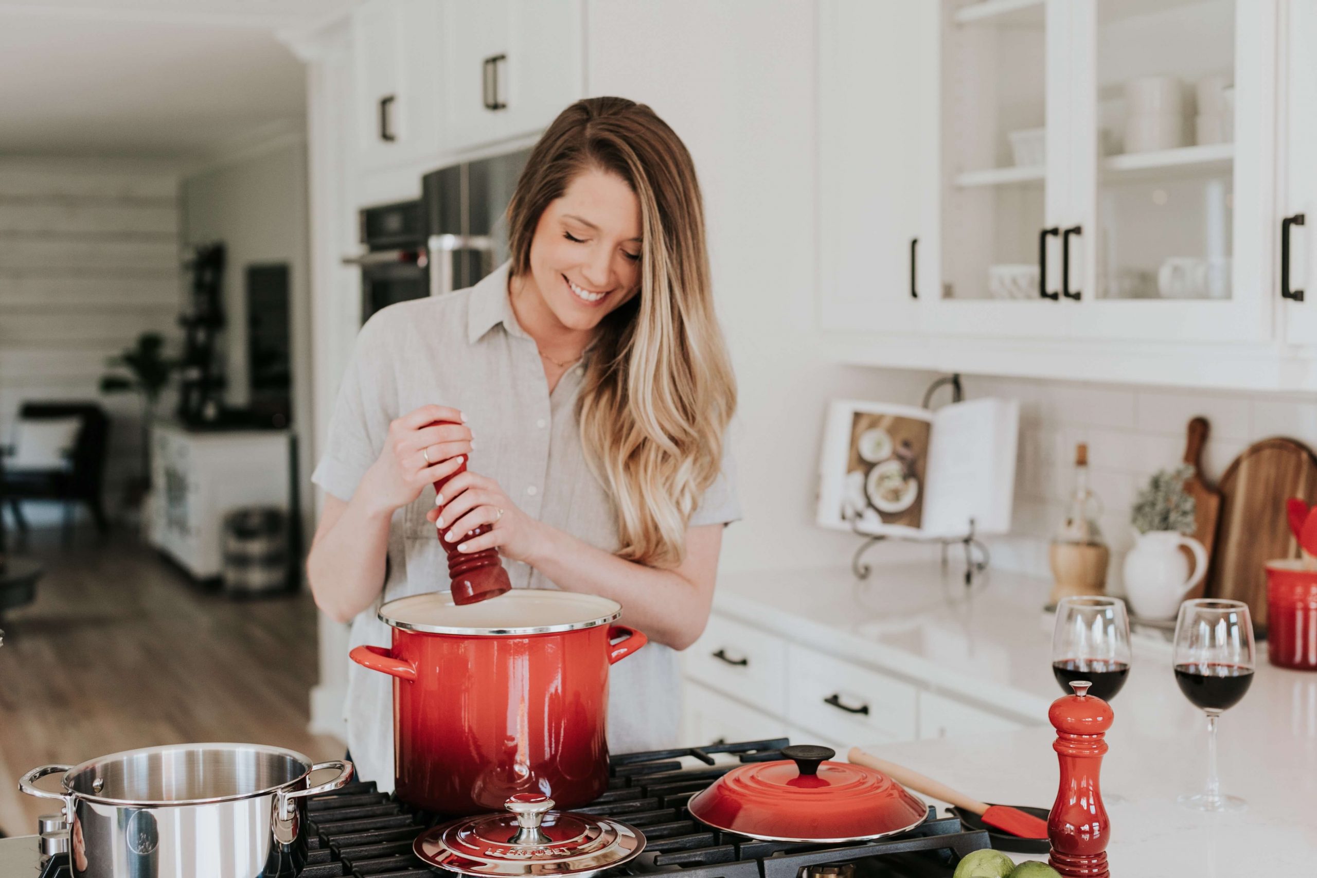 A smiling woman adds ingredients to a red pot on the stove in her bright kitchen, crafting a delightful kid-friendly meal. There are various kitchen items around, including wine glasses and a pepper grinder.