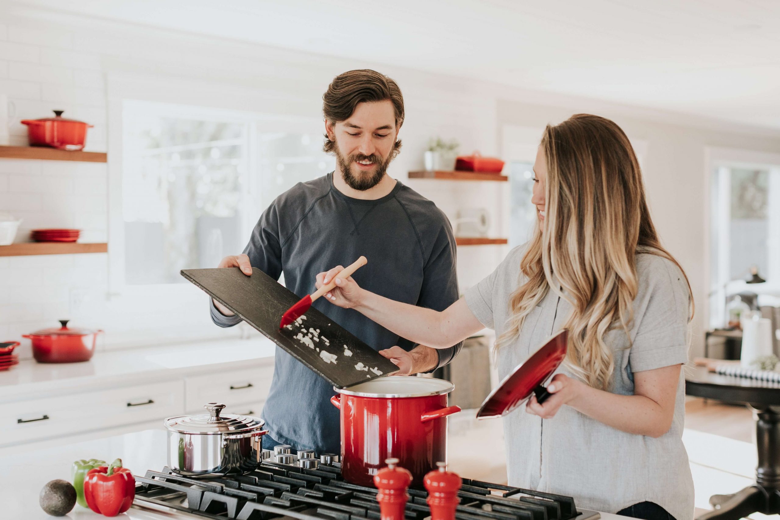 A man and a woman are cooking in a bright kitchen, preparing summer lunch ideas for kids. The man holds a cutting board with chopped onions, while the woman stirs a red pot on the stove, surrounded by a white backsplash and several red kitchenware items.