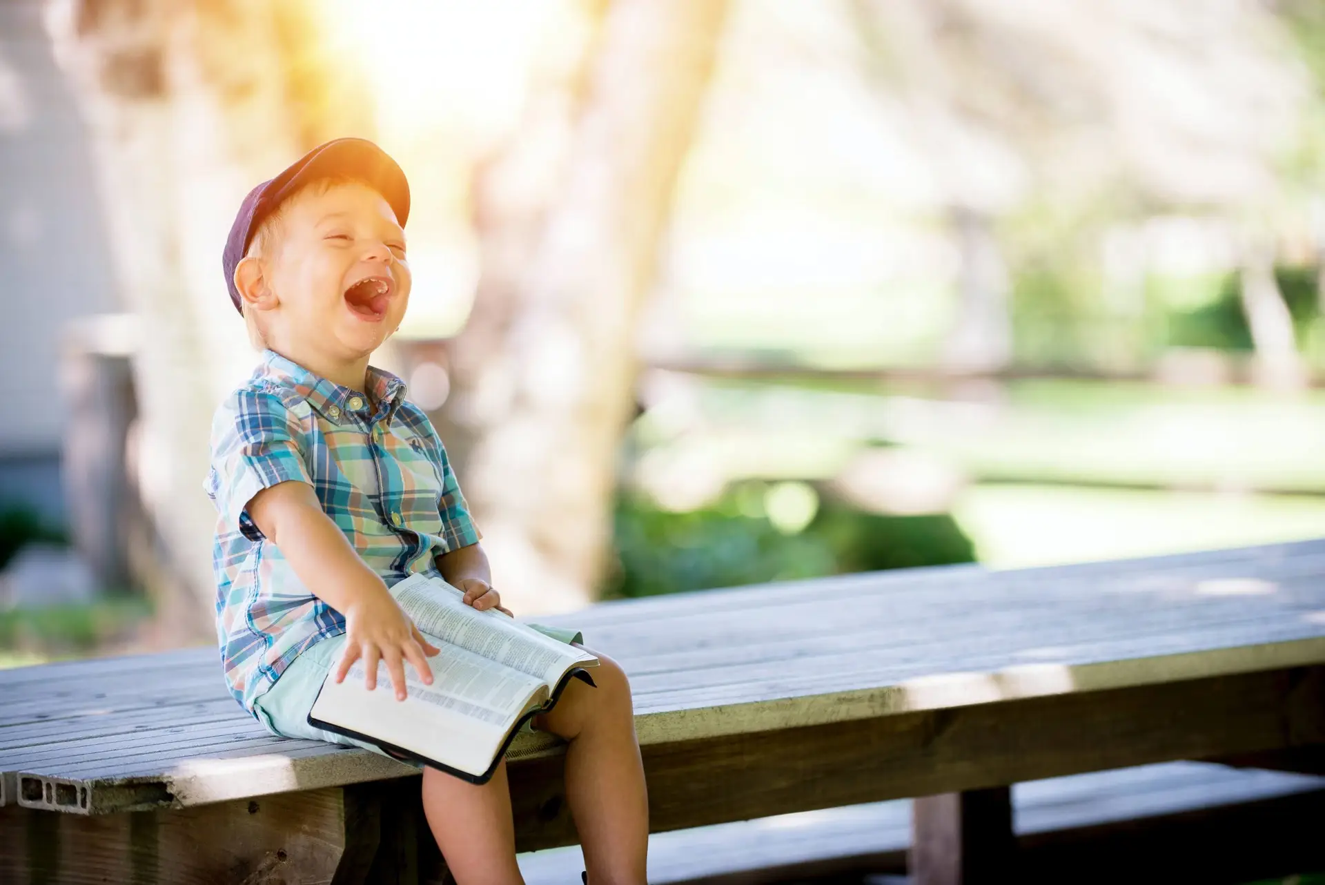 A young child in a plaid shirt and cap sits on a wooden bench outdoors, laughing joyfully with an interactive book on their lap. The background is a sunlit park with blurred trees and greenery.