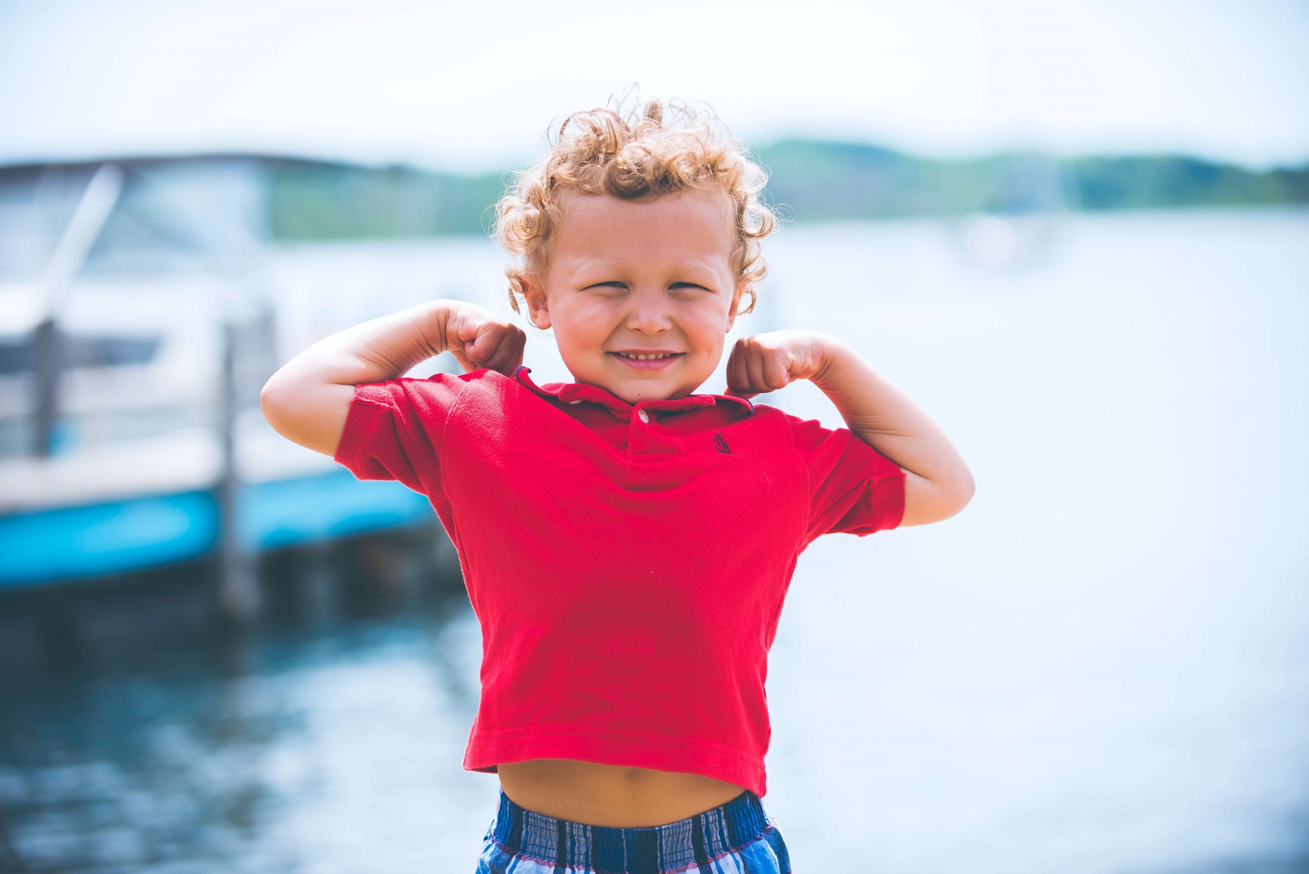 A young child with curly hair smiles and flexes their arms, confidently wearing kids' safety goggles along with a red shirt and plaid shorts. They stand by a waterfront, where a blurred boat drifts under the clear sky.