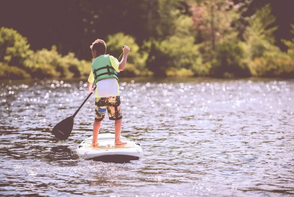 A child wearing a green life jacket stands confidently on a paddleboard, embodying the essence of summer sports. The sunlit lake sparkles with energy as the young adventurer holds a paddle in one hand, against a backdrop of lush greenery.