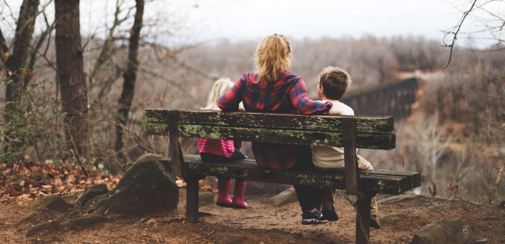 A woman and two children sit on a weathered bench, connecting with kids as they overlook a scenic forest landscape. The group, dressed in casual attire with the children wearing jackets, enjoys the serene autumnal atmosphere created by bare trees and a misty horizon.