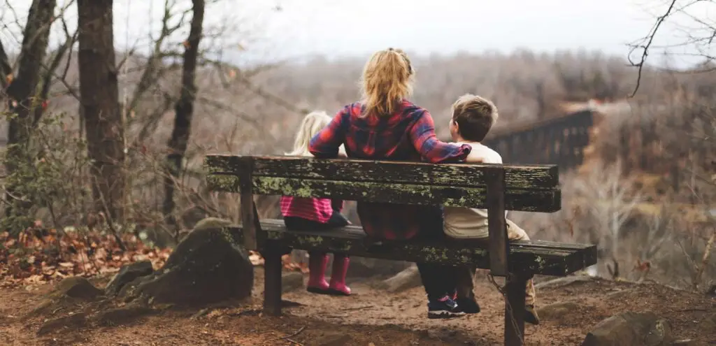 A woman and two children sit on a weathered bench, connecting with kids as they overlook a scenic forest landscape. The group, dressed in casual attire with the children wearing jackets, enjoys the serene autumnal atmosphere created by bare trees and a misty horizon.