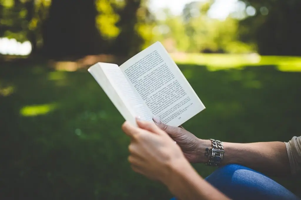 A person is sitting outdoors on the grass, immersed in a book about ADHD for parents. The photo captures a sunny day in the park, with them wearing blue jeans and a watch. In the background, vibrant green trees stand against a blurred, sunlit landscape.