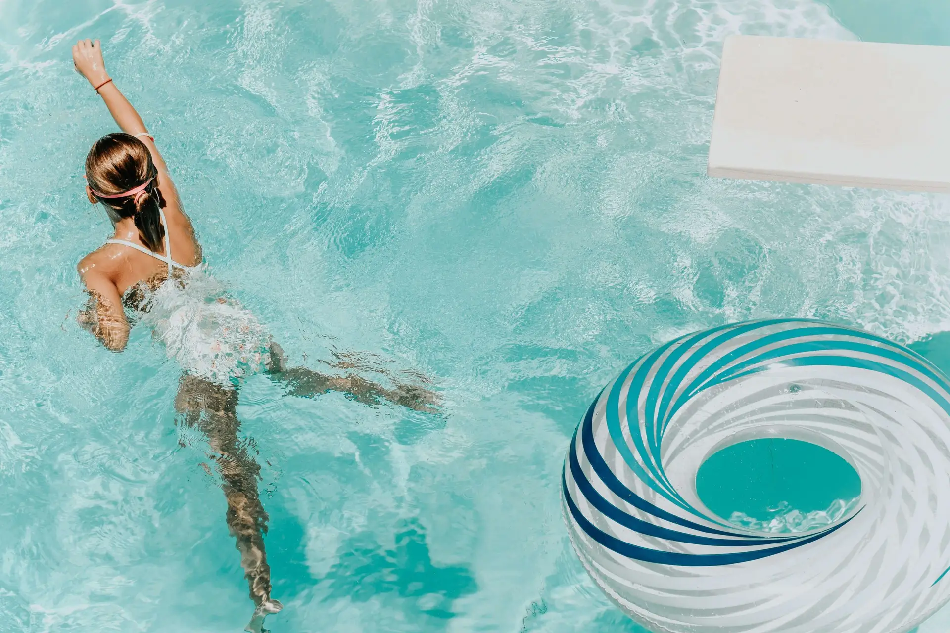 A girl in a white swimsuit gracefully swims through the clear blue pool, ticking another adventure off her summer bucket list for kids. She sports a pink headband, her right arm rising elegantly beside a blue and white striped inflatable ring that floats nearby.