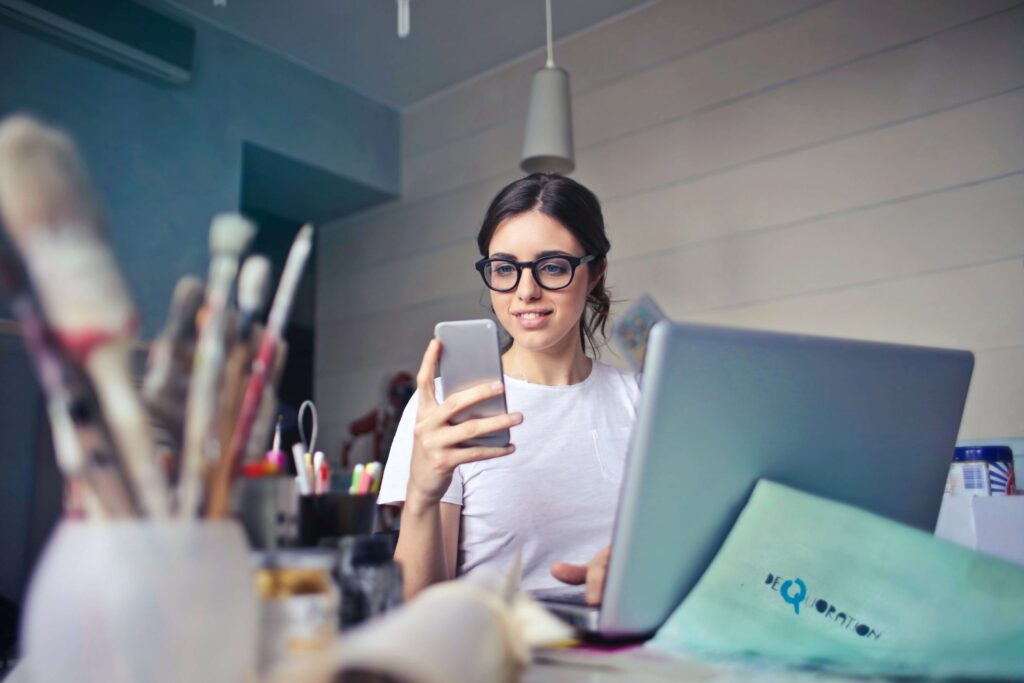 A woman wearing glasses sits at a desk with her smartphone and open laptop, surrounded by art supplies and brushes, creating in this inspiring workspace. As she researches the best free parental control apps, creativity and innovation blend seamlessly into her daily routine.