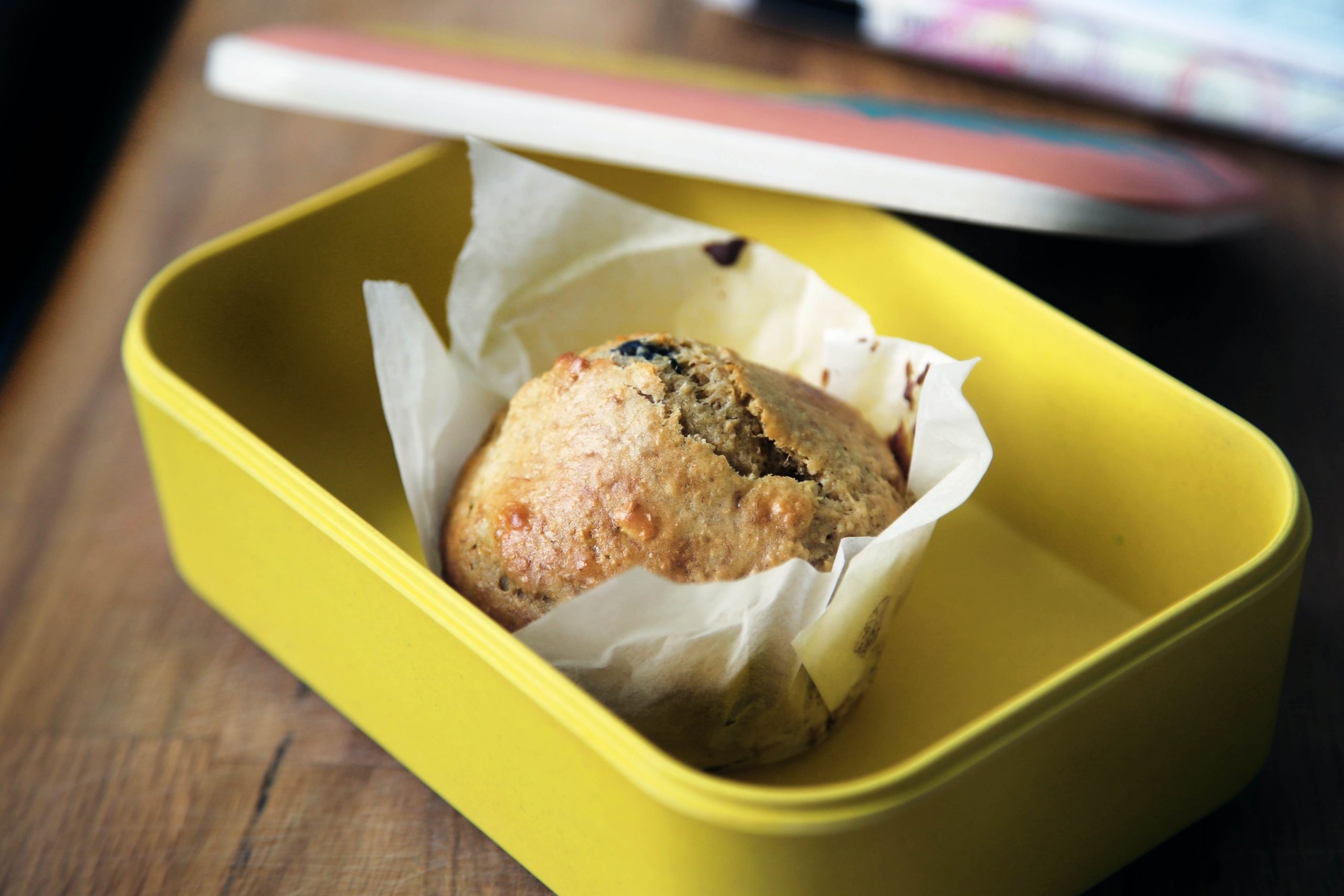 A muffin, nestled in parchment paper, rests inside a yellow rectangular snack container for kids on a wooden surface. In the background, a book with a colorful cover peeks out playfully.