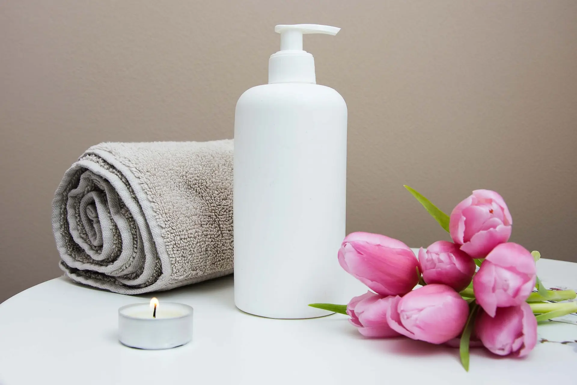 A white pump bottle with lotion sits on a white surface beside a rolled gray towel and a small lit candle, perfect for a kids spa party. A bunch of pink tulips lies nearby, while the beige wall background creates a serene spa-like setting.