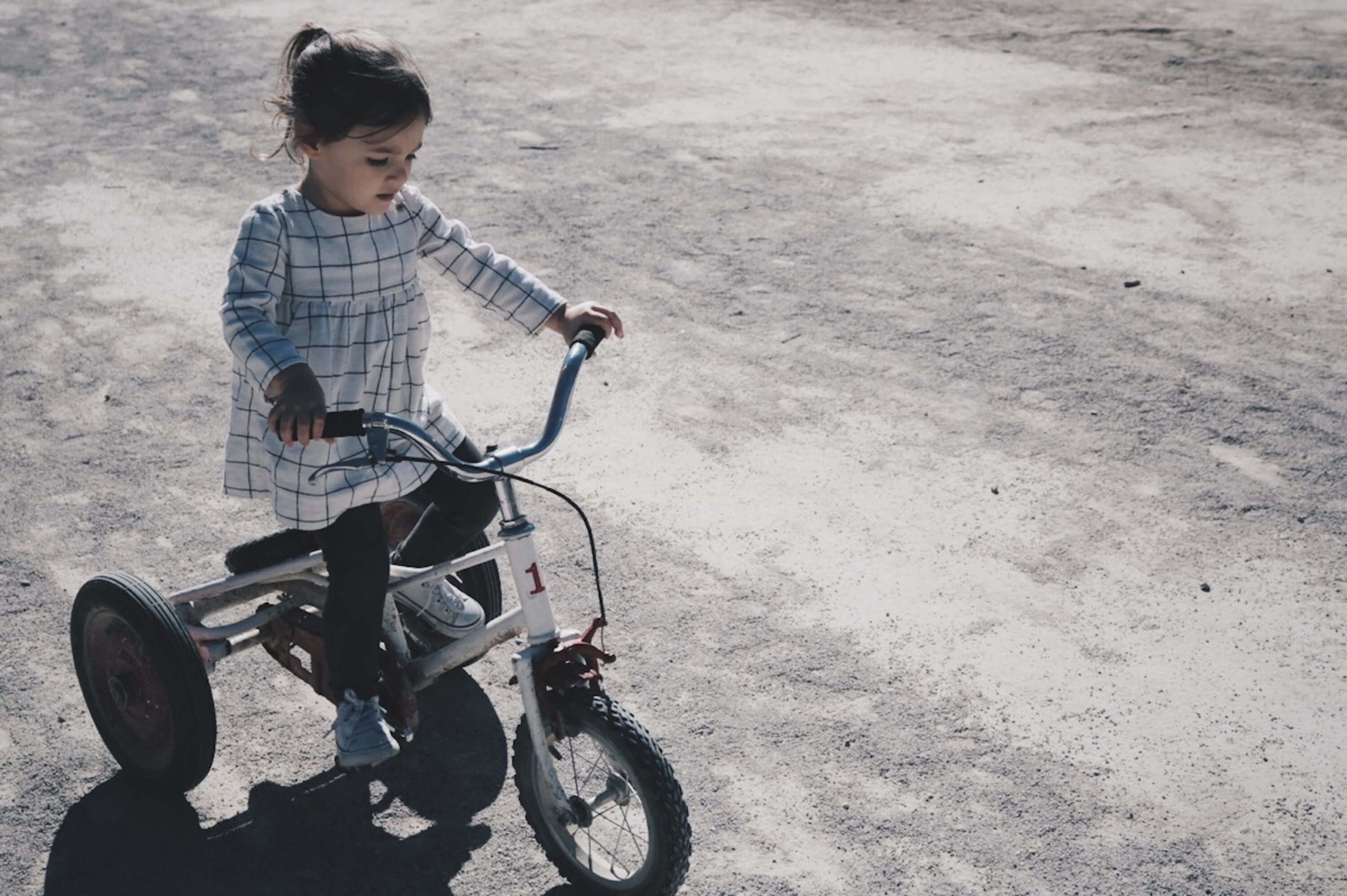 A young child in a plaid dress rides a tricycle with training wheels on a sunlit dirt path. Shadows dance on the ground as the bright sunlight illuminates their focused expression—a charming moment in learning how to teach kids to ride a bike.