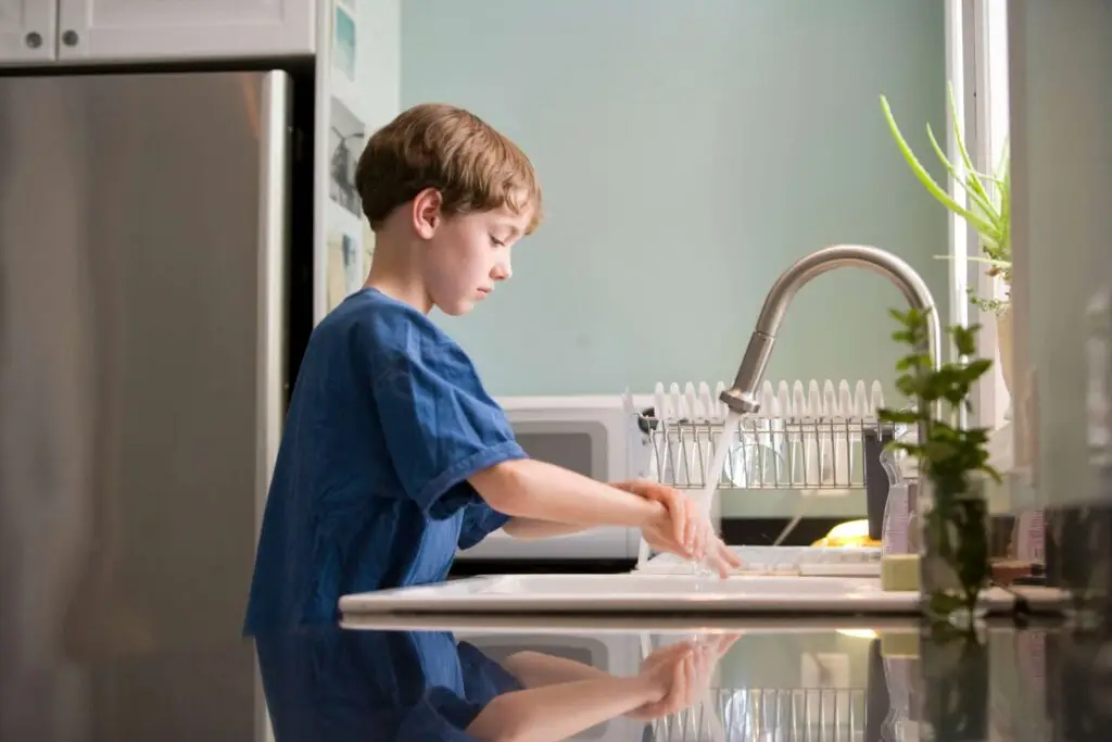 A young boy with short brown hair, wearing a blue shirt, stands at a kitchen sink washing his hands under the running faucet. Sunlight streams through a window, highlighting the dish rack and plant on the counter, reminders of safe kitchen habits like knife safety for kids.