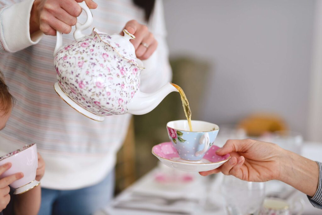 A person pours tea from a floral teapot into a delicate, patterned teacup on a saucer held by another person. Nearby, children hold similar teacups near their faces. The scene evokes the charm of a cozy kids' tea party gathering.