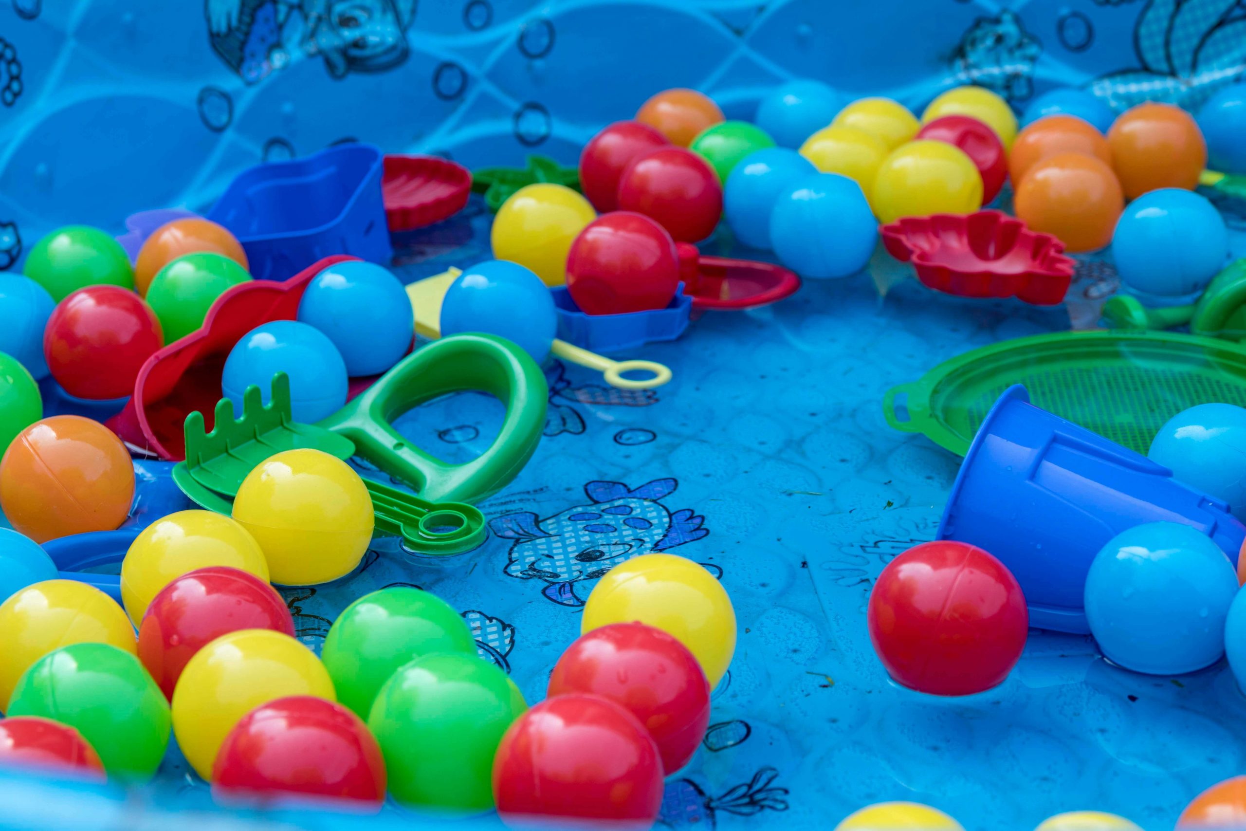 A blue kiddie pool filled with vibrant plastic balls in red, yellow, blue, and green sits invitingly. Surrounding it are playful plastic toys like a green crab claw clapper and a blue bucket. For added kiddie pool safety, the pool features a charming fish pattern on the bottom.