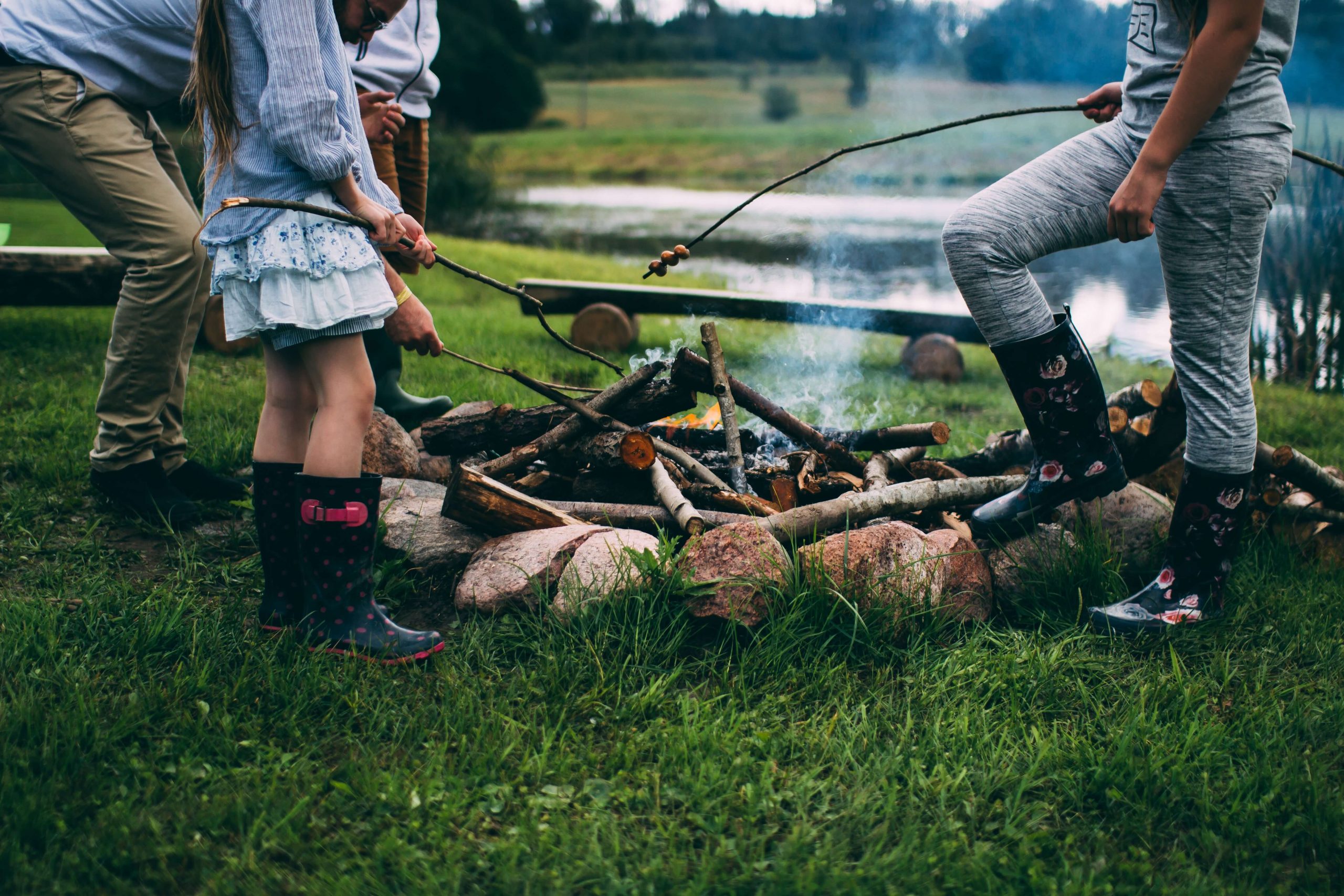 People are standing around a campfire, toasting sticks with marshmallows, enjoying one of the classic camping activities for kids. They are wearing casual clothes and rubber boots. The setting is outdoors, near a body of water, with grassy surroundings and logs encircling the fire.