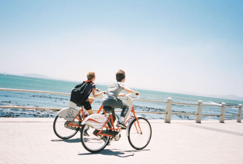 Two people are riding bicycles along a seaside path under a clear blue sky. The ocean is visible to the left, with waves gently crashing onto the rocky shore. Alongside them, an electric bike for kids zips by, adding to the enjoyment of this sunny day adventure.