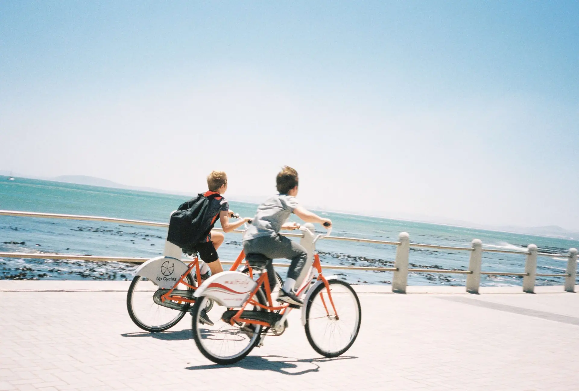 Two people are riding bicycles along a seaside path under a clear blue sky. The ocean is visible to the left, with waves gently crashing onto the rocky shore. Alongside them, an electric bike for kids zips by, adding to the enjoyment of this sunny day adventure.