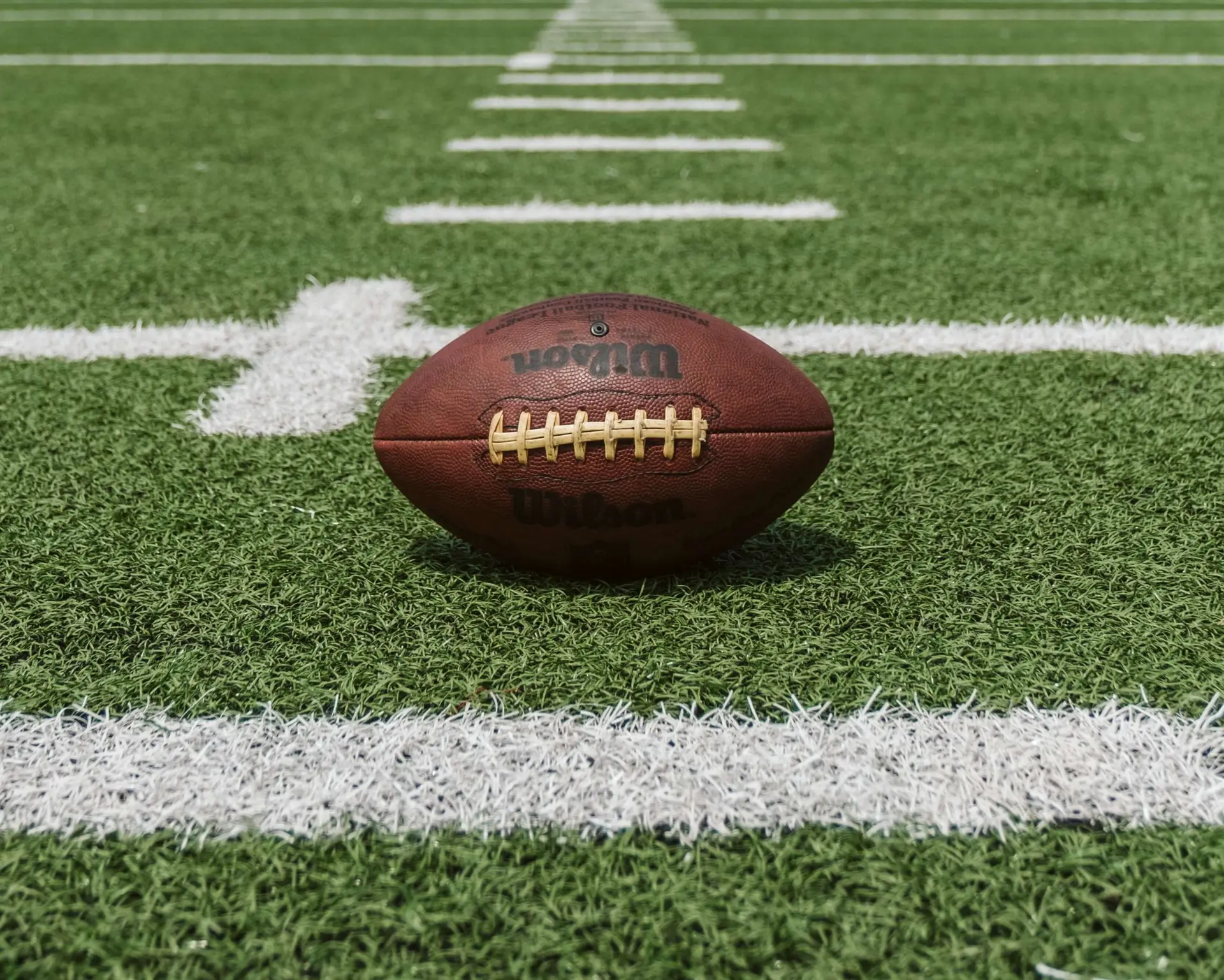 A football rests on a green artificial turf field, centered on a white yard line. The focus is on the football, perfect for setting up football drills for kids, with the horizontal lines of the field leading into the distance.