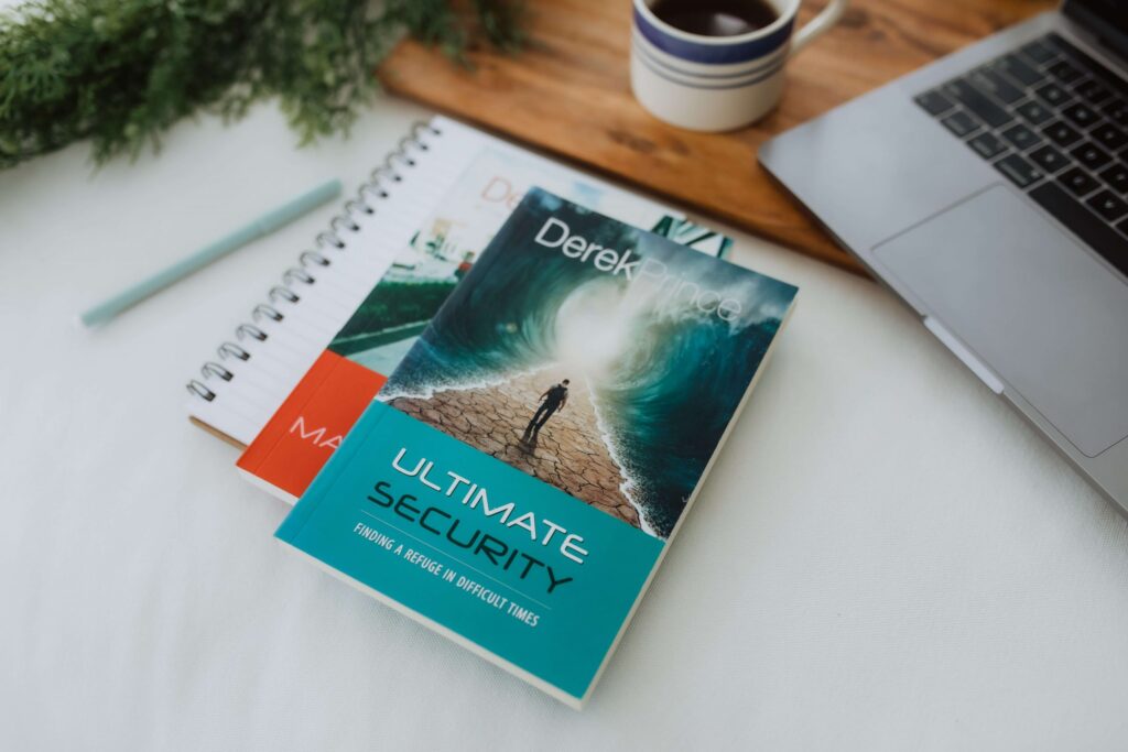 A teal book titled Ultimate Security by Derek Prince, a well-regarded Christian book about parenting, rests on a white surface next to an open notebook and pen. Nearby, a laptop and cup of coffee sit on a wooden tray, with green foliage partially visible in the corner.