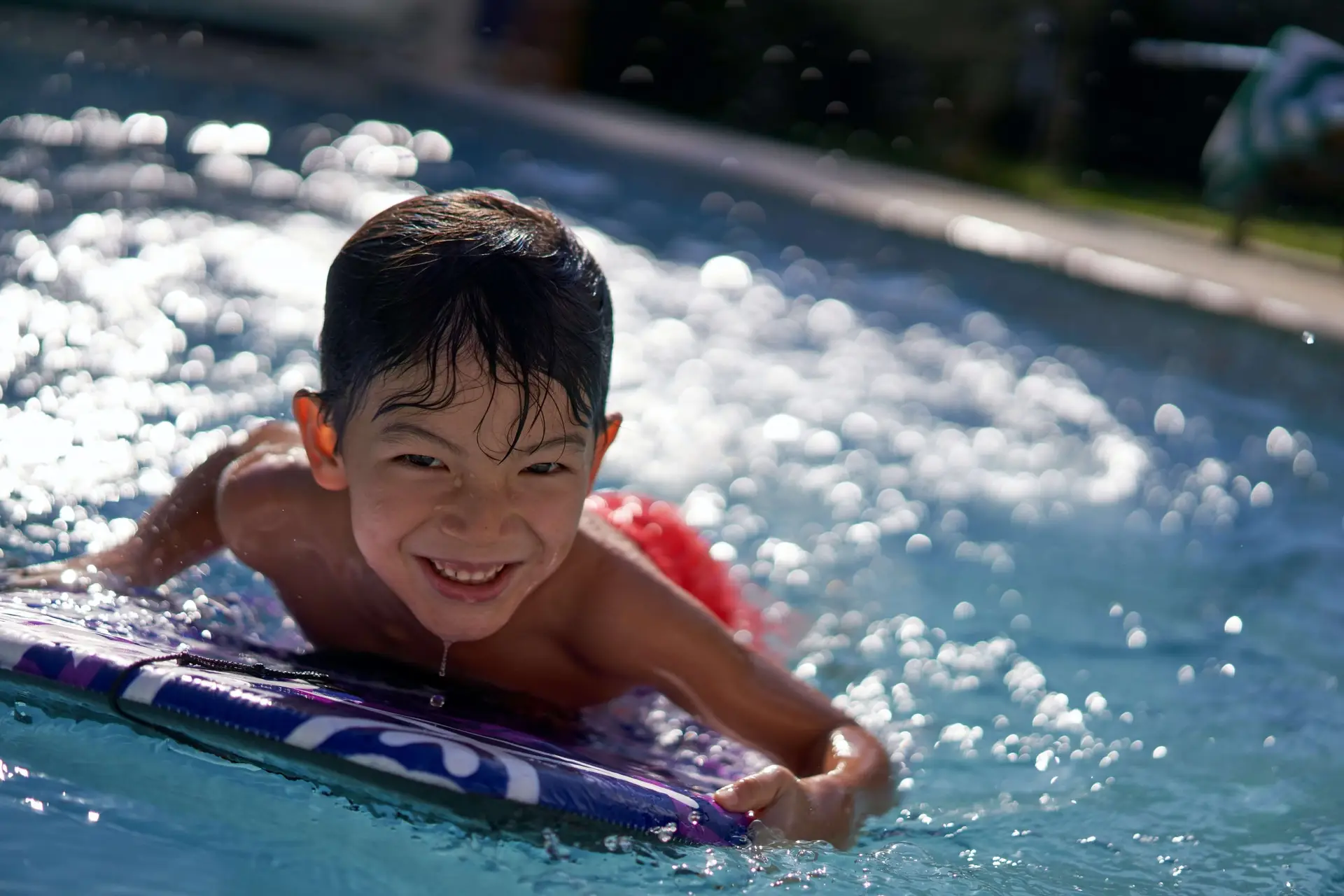 A smiling child with wet hair is lying on a boogie board in a swimming pool, embodying the joy of learning how to swim. They're wearing red swim trunks and appear to be having fun as water splashes around them. The sun reflects off the water, creating a bright, cheerful scene.