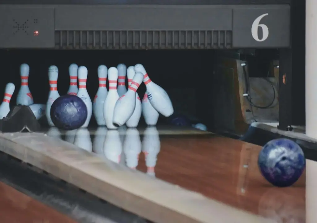 A bowling ball strikes a set of white pins with red stripes on a polished wooden lane. Another blue ball is visible in the foreground, adding excitement to the bowling party for kids. The number 6 is displayed above the pin area, marking the fun-filled action underway.