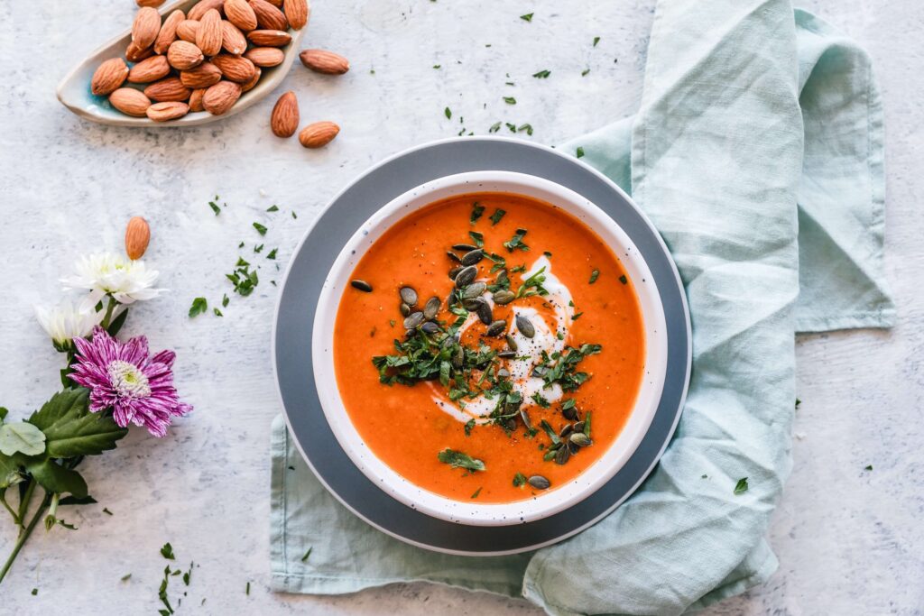 A bowl of kid-friendly tomato soup garnished with cream, pumpkin seeds, and chopped herbs sits on a light gray surface with a pale blue cloth. Nearby, a small dish of almonds and a few flowers add charm to the scene.