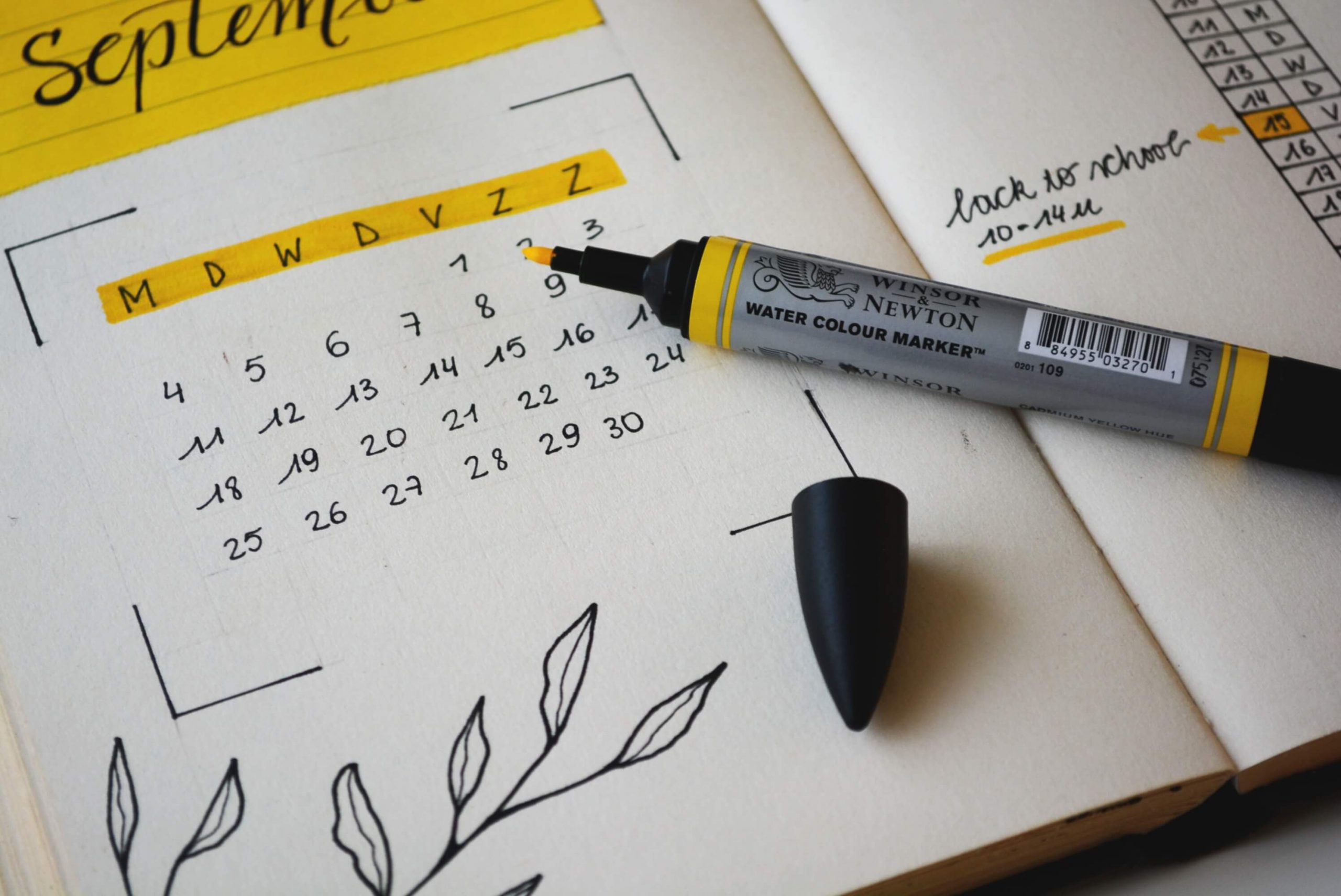 A journal features an interactive September calendar for kids, hand-drawn with black and yellow accents. A black cap is detached from a yellow Winsor & Newton watercolor marker lying on the page. A note on the right reads back to school, with arrows pointing to the 10th-14th.