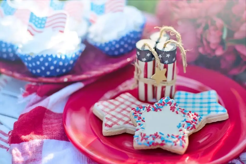 A festive display includes 4th of July cookies shaped like stars, decorated with red, white, and blue sprinkles and patterns. A candle resembling a firecracker and cupcakes in patriotic wrappers complement the scene, set on a red plate with a checkered cloth.