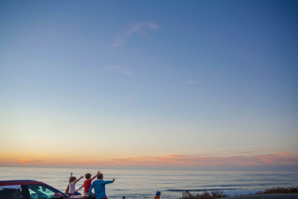 A group of people stands by the ocean during sunset, gesturing and pointing at the sky. The horizon is painted with soft pink and orange hues, perfect for capturing car activities for kids. A vehicle is parked next to them while the vast, colorful sky dominates the image.