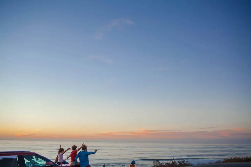 A group of people stands by the ocean during sunset, gesturing and pointing at the sky. The horizon is painted with soft pink and orange hues, perfect for capturing car activities for kids. A vehicle is parked next to them while the vast, colorful sky dominates the image.
