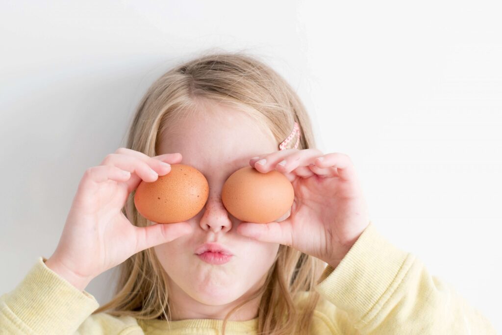 A child, in a light yellow shirt, holds two brown eggs up to their eyes, playfully imitating glasses with a puckered expression. These protein-packed snacks for kids create a fun and healthy moment against the plain white background.