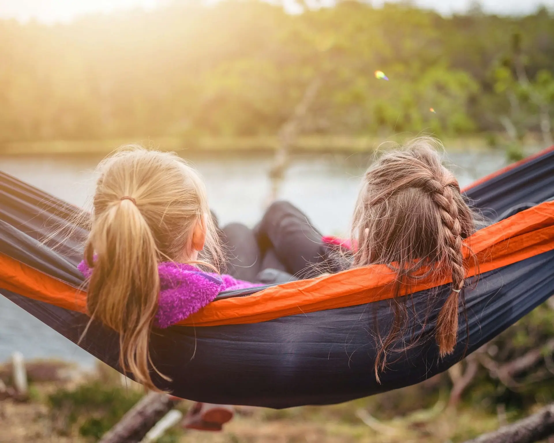 Two children with braided hair relax in an orange and black hammock, each holding books about making friends, as they overlook a serene body of water surrounded by lush greenery. The sun casts a warm glow, creating a peaceful outdoor scene.