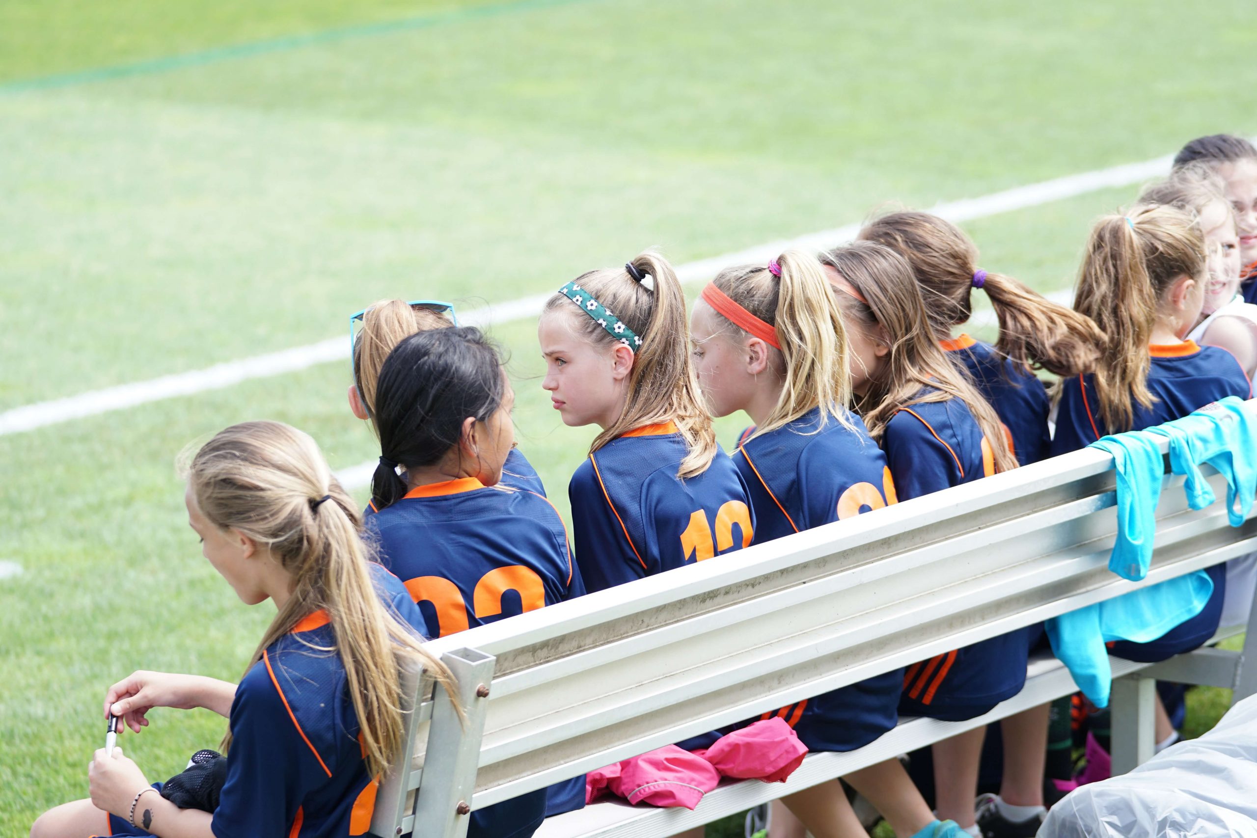 A group of young girls wearing navy sports jerseys with orange numbers are sitting on a bench by a soccer field, their kids' prescription sports glasses glinting in the sun. Some are chatting, while others look onto the lush green field. The team seems to be taking a break.