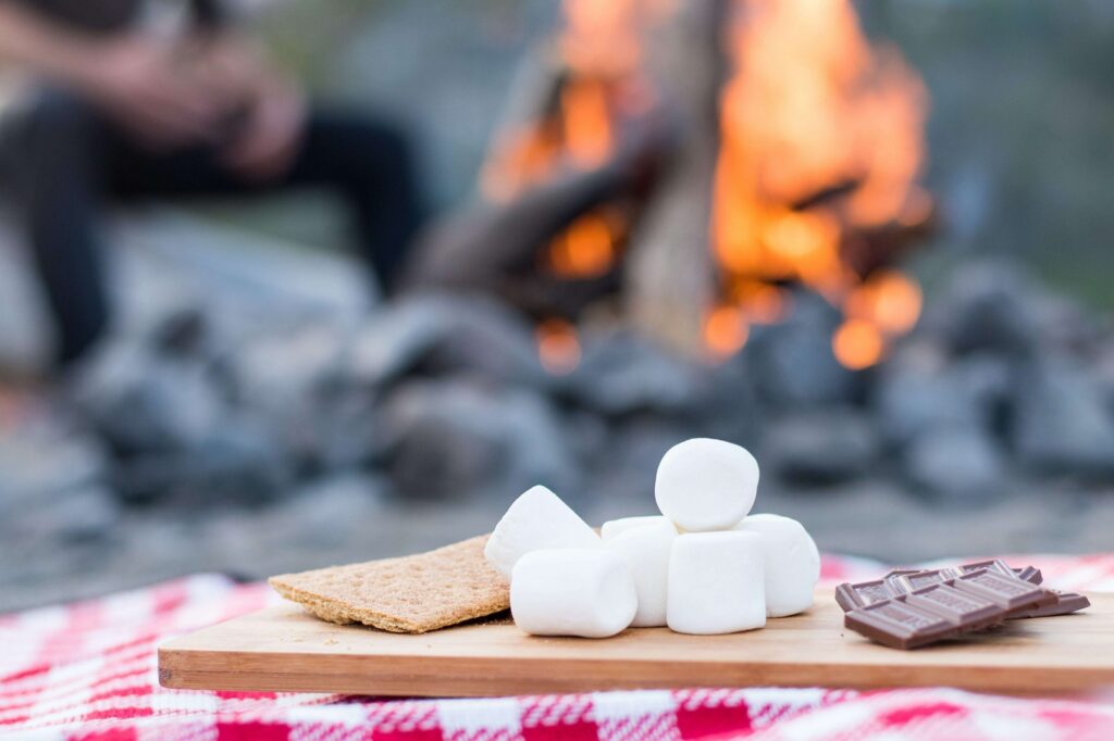 Ingredients for s'mores are artfully arranged on a wooden board: graham crackers, marshmallows, and chocolate. A picnic blanket and a campfire in the background inspire cozy s'mores ideas for your next outdoor adventure.