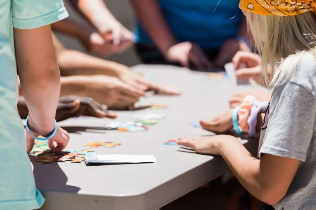 People gathered around a table outdoors, working on a jigsaw puzzle. Sunlight casts shadows on the group as they focus on assembling colorful pieces. A child with a yellow bandana enthusiastically shares sports trivia for kids while helping connect the pieces together.